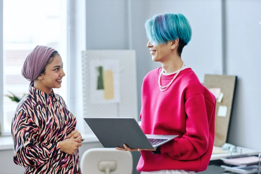 Two gen Z young people chatting standing in office and holding laptop