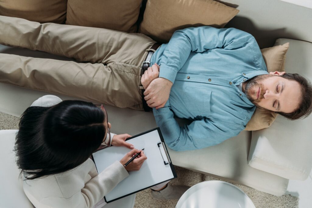 overhead view of psychotherapist writing on clipboard and male patient with closed eyes lying on